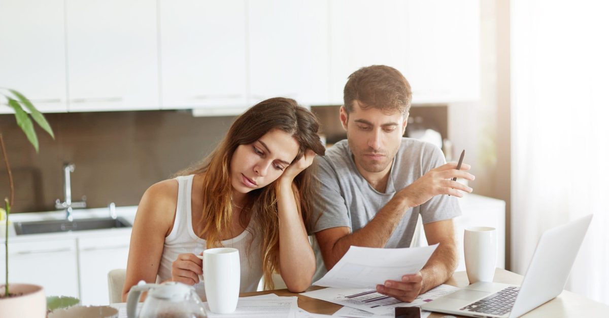 People, money and finances concept. Young tired Caucasian woman leaning at her hand and drinking morning coffee while sitting near her husband at kitchen table, working with documents and laptop